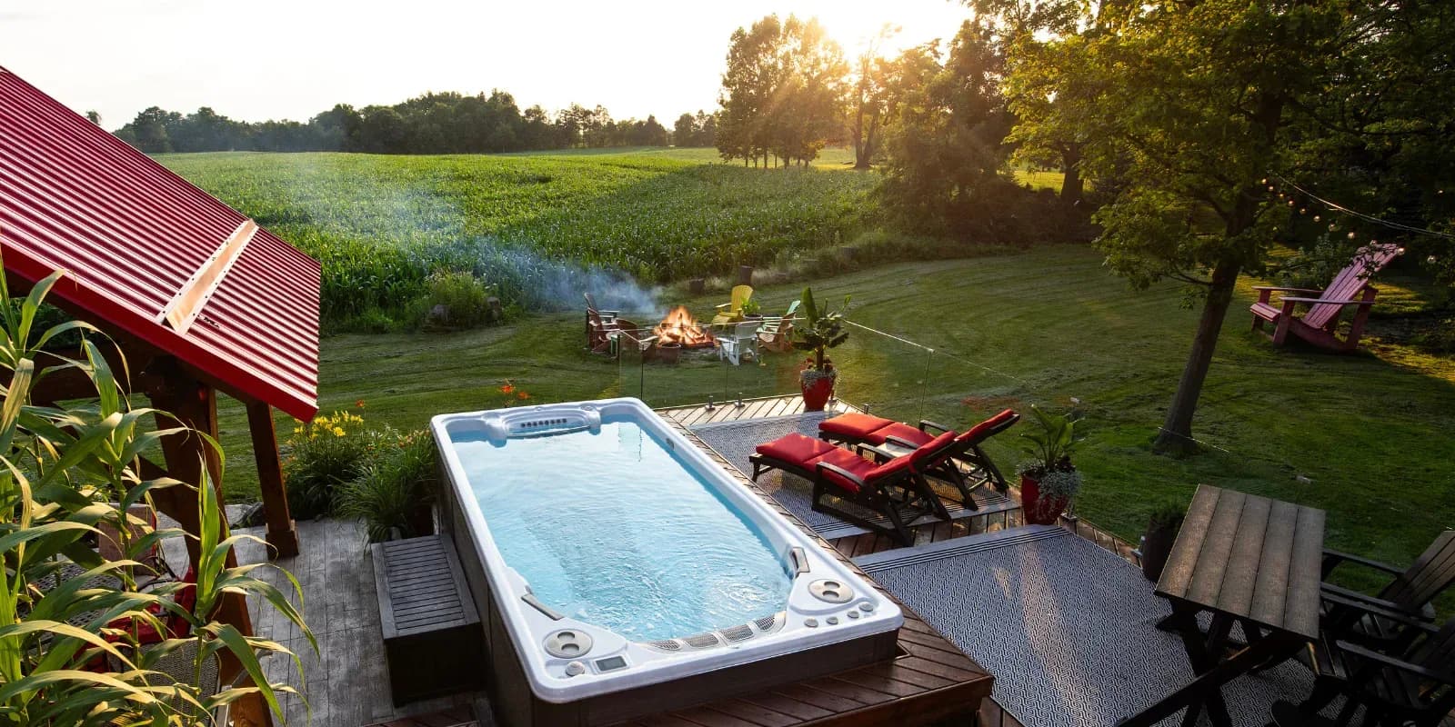 Hydropool swim spa set into a backyard deck at sunset, with red loungers, a fire pit and a cornfield beyond