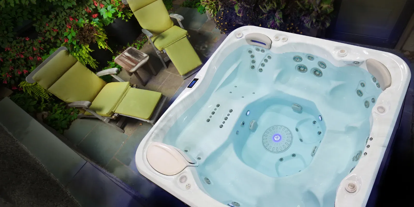 Hydropool hot tub seen from above at night on a stone patio, with two green-cushioned loungers and garden plants beside it